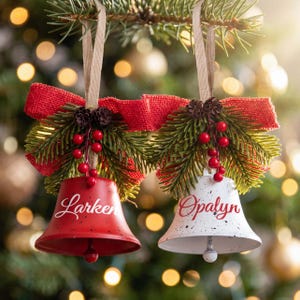 Può includere: Due ornamenti a forma di campana appesi a nastri color cuoio, uno rosso e uno bianco, con macchie nere e i nomi Larken e Opalyn in corsivo. Sono decorati con fiocchi di juta rossa, aghi di pino verdi, piccole pigne e bacche rosse.