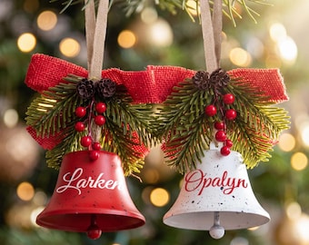 May include: Two bell ornaments hanging from tan ribbons, one red and one white, each featuring black speckles and the names Larken and Opalyn in script. They are decorated with red burlap bows, green pine needles, small pinecones, and red berries.