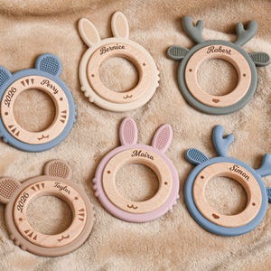 May include: Six baby teething toys in various pastel colors, including blue, pink, and beige. Each toy features a wooden ring with a silicone animal shape, such as a bear, bunny, or deer. Names and the year 2026 are engraved on the wooden rings.