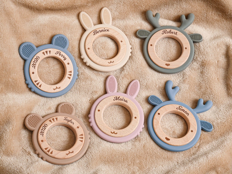 May include: Six baby teething toys in various pastel colors, including blue, pink, and beige. Each toy features a wooden ring with a silicone animal shape, such as a bear, bunny, or deer. Names and the year 2026 are engraved on the wooden rings.