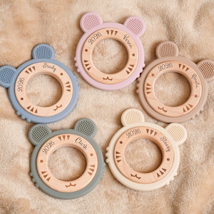 May include: Five baby teething toys in various pastel colors, each with a wooden center and a silicone outer ring shaped like a bear's head. Each toy has a name and the year 2026 engraved on it.