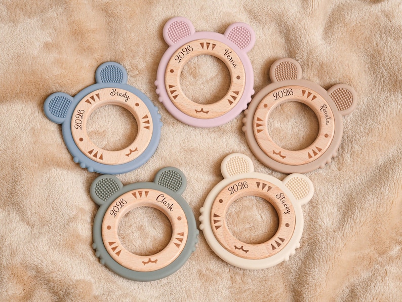 May include: Five baby teething toys in various pastel colors, each with a wooden center and a silicone outer ring shaped like a bear's head. Each toy has a name and the year 2026 engraved on it.