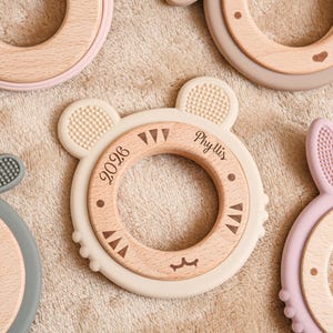 May include: A collection of baby teethers in various colors and shapes. The teethers feature a wooden ring with a silicone outer ring in the shape of a bear, bunny, and other designs. The bear teether has the name "Phyllis" and the year "2026" engraved on it.