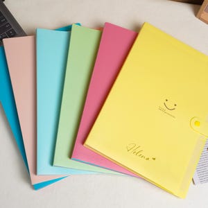 May include: A set of six rectangular document folders in assorted pastel colours including yellow, pink, light green, light blue, peach, and dark blue. The yellow folder features a snap button closure, a printed smiley face, and the name Helena in script.