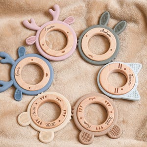 May include: A collection of six baby teething toys in various pastel colors, including pink, blue, and beige. Each toy features a wooden center ring and a silicone outer ring with different animal-themed shapes, such as antlers and bunny ears. Names are engraved on the wooden rings.