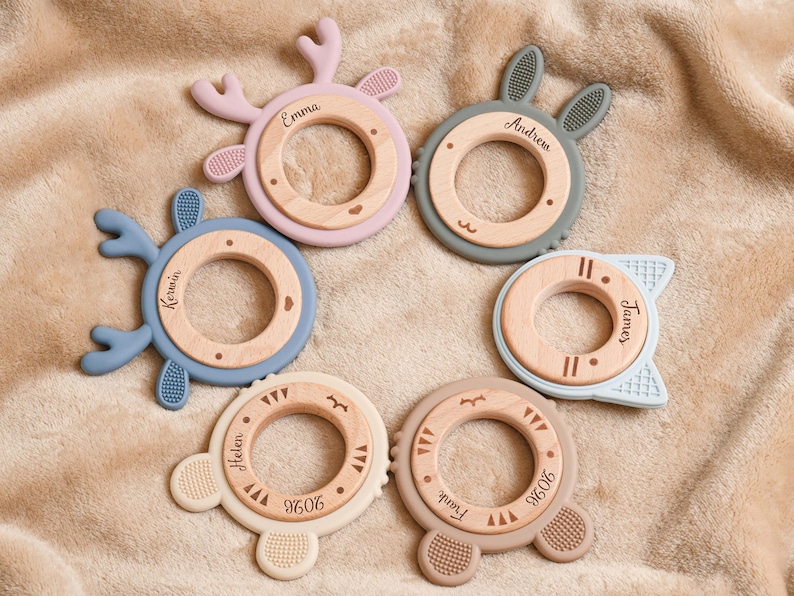 May include: A collection of six baby teething toys in various pastel colors, including pink, blue, and beige. Each toy features a wooden center ring and a silicone outer ring with different animal-themed shapes, such as antlers and bunny ears. Names are engraved on the wooden rings.