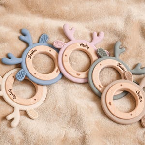 May include: Five baby teething rings in various pastel colors, each with a wooden center and silicone antlers. The names "Wesley", "Justin", "Molly", "Fruma", and "Cecilia" are printed on the wooden rings.