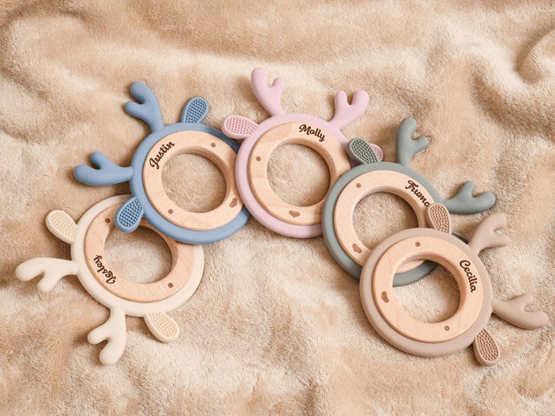 May include: Five baby teething rings in various pastel colors, each with a wooden center and silicone antlers. The names "Wesley", "Justin", "Molly", "Fruma", and "Cecilia" are printed on the wooden rings.