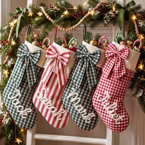 May include: Four Christmas stockings hanging in a row, each featuring a large fabric bow at the top and a name embroidered in white script: Jack, Sarah, Mark, and Juliet. The stockings alternate between green gingham, red stripes, and red gingham patterns.