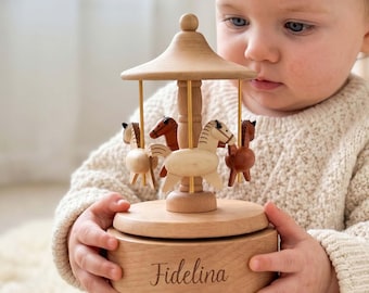 May include: A light-colored wooden carousel music box featuring four small carved horses in varying shades of brown and tan. The base of the circular wooden toy is engraved with the name Fidelina in a cursive script font, with gold-toned metal rods.