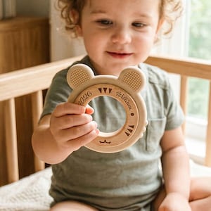 May include: A wooden baby teether in the shape of a bear with the name "William" engraved on it. The teether has a circular design with textured ears and a smooth, natural wood finish. The text "0026" is also engraved on the teether.