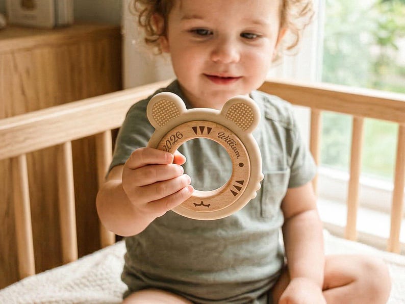 May include: A wooden baby teether in the shape of a bear with the name "William" engraved on it. The teether has a circular design with textured ears and a smooth, natural wood finish. The text "0026" is also engraved on the teether.