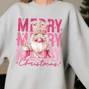 以下が含まれることがあります： ピンク色のクリスマス柄がプリントされたライトグレーのスウェットシャツ。王冠とメガネをかけたくるみ割り人形と、ピンク色の「MERRY CHRISTMAS」の文字がデザインされています。