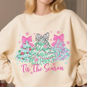 May include: Cream-colored sweatshirt featuring a festive design of three Christmas trees with pink bows and a black and white polka dot bow. The phrase "Tis The Season" is printed in pink script.
