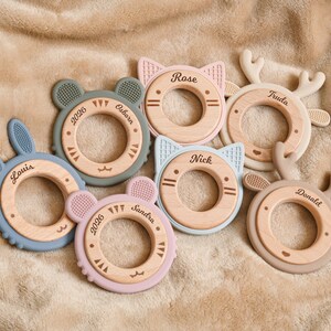May include: A collection of baby teethers in various pastel colors, including blue, pink, and green. Each teether features a wooden ring with a silicone animal shape, such as a bunny, cat, bear, or deer. Names and dates are printed on the wooden rings.