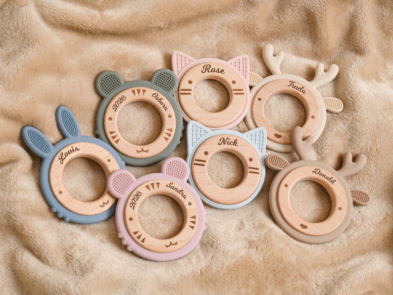 May include: A collection of baby teethers in various pastel colors, including blue, pink, and green. Each teether features a wooden ring with a silicone animal shape, such as a bunny, cat, bear, or deer. Names and dates are printed on the wooden rings.