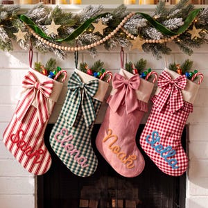 May include: Four Christmas stockings hanging in a row, each featuring a large fabric bow at the top and a name embroidered in cursive script. The stockings display red and white stripes, green and white plaid, solid pink, and red and white gingham patterns.