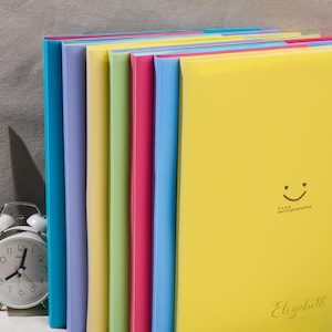 May include: A set of seven colorful plastic document folders arranged in a row. The folders are blue, purple, yellow, green, pink, light blue, and yellow. The front yellow folder features a black smiley face, the text Keep smilingeverytime, and Elizabeth.