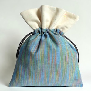 May include: A drawstring bag with a blue, green, and yellow striped pattern. The top of the bag is a cream color, and the drawstring is black. The bag is made of fabric and is likely used for storage or carrying items.