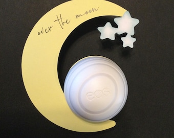 May include: A white EOS lip balm sphere with the word "EOS" printed on it, sitting inside a yellow crescent moon cutout with the text "over the moon" written on it. There are three blue and white star cutouts above the moon.