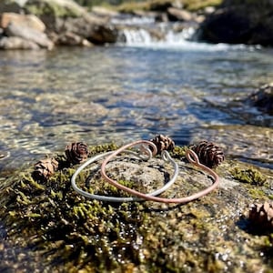 May include: Two handcrafted wire bracelets, one silver and one copper, rest on a moss-covered rock. Several pine cones are scattered around the bracelets. The background shows a flowing stream and rocks.