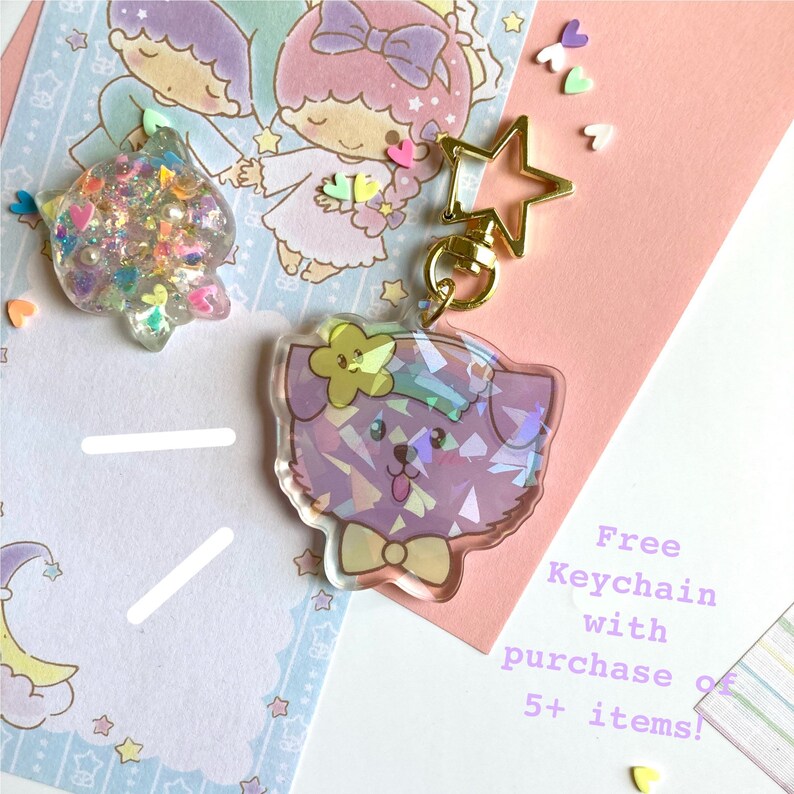 Pu&ograve; includere: Un portachiavi iridescente viola con un cane che indossa un papillon e un fiore sulla testa. Il portachiavi &egrave; attaccato a un portachiavi dorato a forma di stella. Il testo "Free Keychain with purchase of 5+ items!" &egrave; visibile nell'immagine.