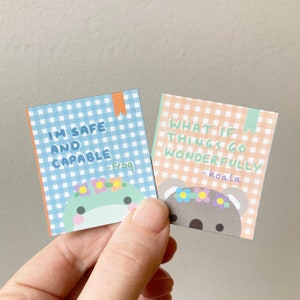 Pu&ograve; includere: Due carte colorate con motivi a quadri vichy e illustrazioni di animali carini. La carta blu dice "Sono al sicuro e sono capace -rana" e la carta arancione dice "E se le cose andassero meravigliosamente bene -koala".