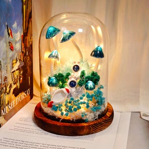 Könnte beinhalten: Dekorative Glaskuppel mit Holzsockel, die eine Miniatur-Fantasieszene zeigt. Sie enthält leuchtende Pilze, winzige Astronautenfiguren und eine kleine Rakete. Die Szene wird von Lichterketten beleuchtet, was eine magische Atmosphäre schafft.