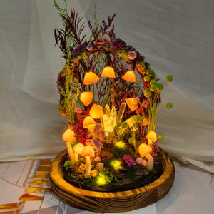 May include: A mushroom-themed terrarium with LED lights. The miniature scene features mushroom figures, crystals, and greenery, all encased in a glass dome. The base is made of wood, and the lights cast a warm glow. The terrarium is a decorative item.