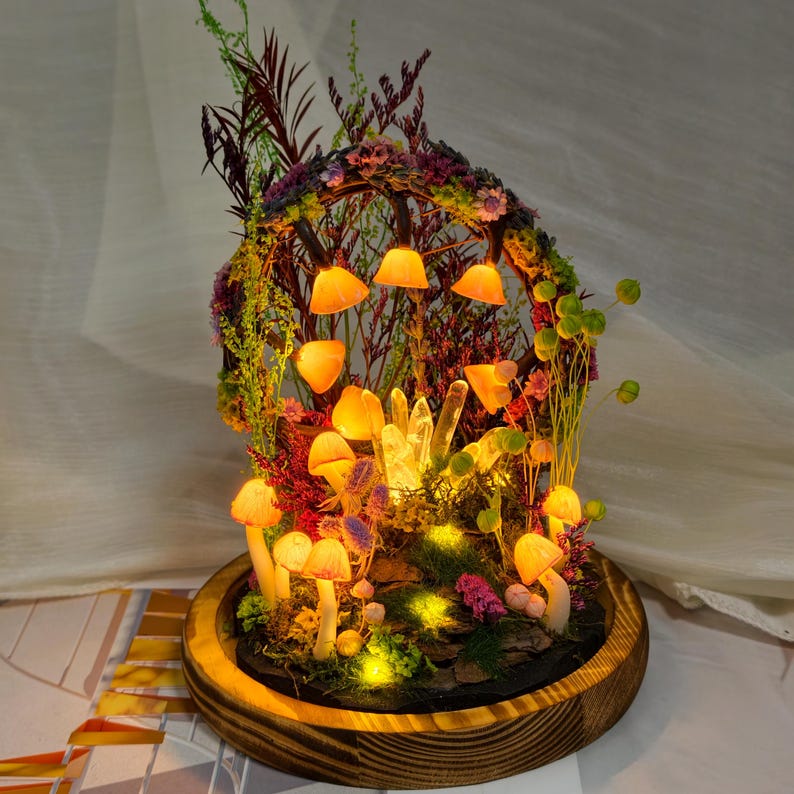 May include: A mushroom-themed terrarium with LED lights. The miniature scene features mushroom figures, crystals, and greenery, all encased in a glass dome. The base is made of wood, and the lights cast a warm glow. The terrarium is a decorative item.