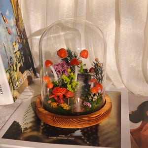 May include: A glass cloche terrarium featuring miniature red mushrooms, purple and yellow flowers, and greenery. The terrarium sits on a wooden base. The scene is reminiscent of a fairy garden. The base is approximately 6 inches in diameter.