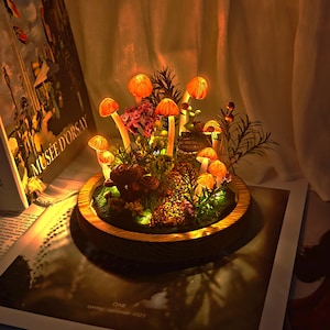 May include: A decorative mushroom terrarium with glowing orange caps and stems, set in a wooden base. The miniature landscape includes greenery and small lights, creating a warm, inviting ambiance. The terrarium is displayed on a book with the text "MUS&Eacute;E D'ORSAY".