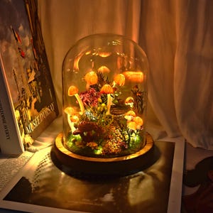 May include: A decorative mushroom-themed lamp encased in a glass dome. The lamp features several illuminated mushrooms with orange caps and white stems, surrounded by greenery and a small path. The base is made of wood, and the lamp is lit with a warm glow.
