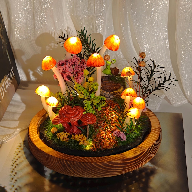 May include: A decorative illuminated mushroom terrarium in a wooden bowl. The terrarium features glowing orange and yellow mushrooms, red mushrooms, and various plants. The mushrooms are lit from within, creating a warm, inviting glow.