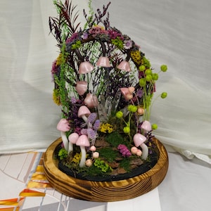 May include: A decorative terrarium with pink mushroom ornaments, quartz crystals, and dried flowers and foliage. The arrangement is set in a wooden base, creating a miniature fairy garden scene.