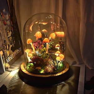 May include: A decorative glass dome featuring illuminated orange mushroom lights, set within a miniature garden with greenery and flowers. The dome sits on a wooden base, creating a whimsical, illuminated display.