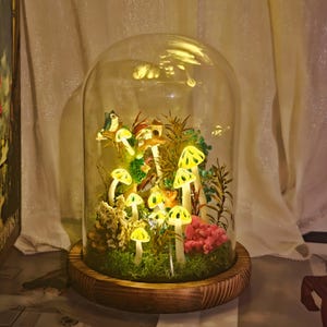 Könnte beinhalten: Eine dekorative Glaskuppel mit Holzsockel, die eine Miniatur-Pilzszene mit Beleuchtung zeigt. Die Szene umfasst leuchtende gelbe Pilze, Miniaturfiguren und Grünzeug, wodurch eine skurrile Darstellung entsteht. Die Kuppel wird von innen beleuchtet und erzeugt einen warmen Schein.