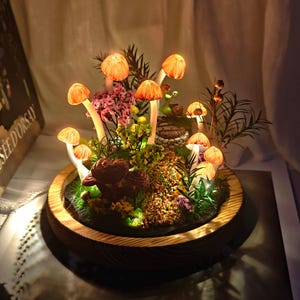 May include: A decorative terrarium featuring illuminated orange mushroom lights, surrounded by greenery, moss, and small flowers. The mushrooms have white stems and are set in a wooden bowl.