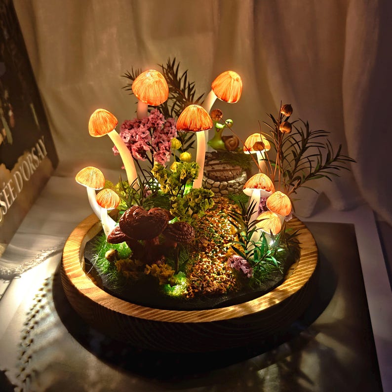May include: A decorative terrarium featuring illuminated orange mushroom lights, surrounded by greenery, moss, and small flowers. The mushrooms have white stems and are set in a wooden bowl.