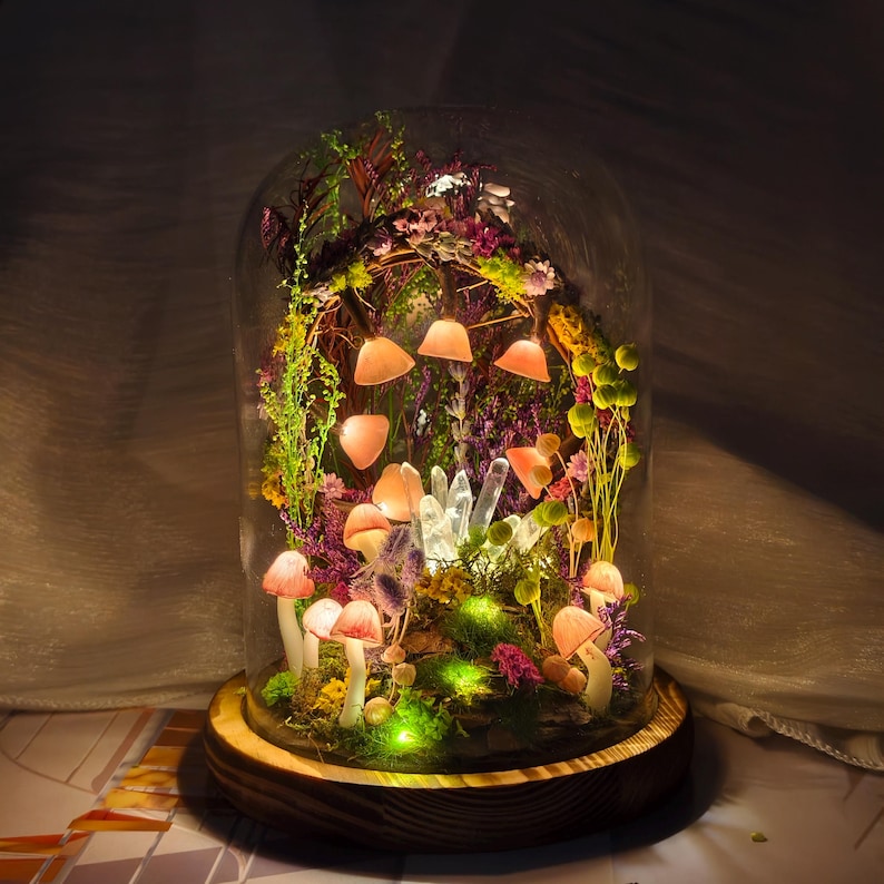 May include: A decorative glass cloche with a wooden base, featuring a miniature illuminated mushroom and floral arrangement. The arrangement includes pink mushrooms, crystals, and various plants, creating a whimsical display. The cloche is lit with warm lights, creating a magical scene.