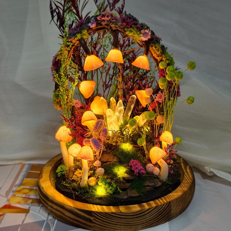 May include: A miniature fairy garden scene featuring glowing mushrooms, crystals, and tiny lights. The display is set within a wooden base and covered by a glass dome, creating an enchanting, illuminated scene.