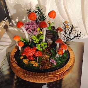 May include: A miniature mushroom garden in a wooden bowl. The garden features red and orange mushrooms with white stems, surrounded by green foliage, purple flowers, and a small stone structure. The bowl has a natural wood grain finish.