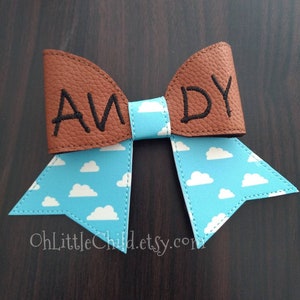 May include: A brown and blue bow with the name "ANDY" embroidered in black. The bow features a light blue fabric with white cloud patterns and is likely a hair accessory or decorative item.