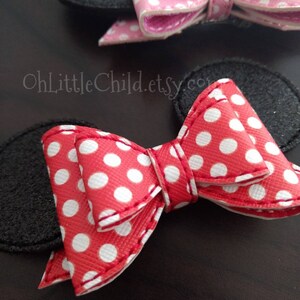 May include: Close-up of two bow-shaped hair accessories. One is red with white polka dots, the other is pink with white polka dots. Both have black felt ear shapes attached. The accessories are on a dark brown surface.