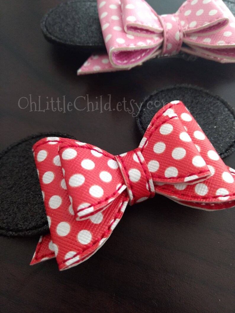 May include: Close-up of two bow-shaped hair accessories. One is red with white polka dots, the other is pink with white polka dots. Both have black felt ear shapes attached. The accessories are on a dark brown surface.