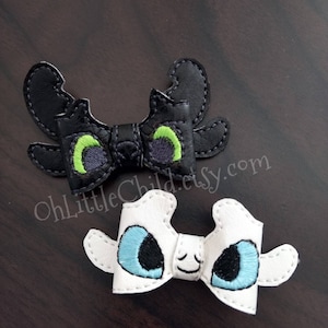 May include: Two embroidered bow hair clips. One is black with green and purple eyes, the other is white with blue and black eyes. The clips are shaped like a dragon's face. The text "OhLittleChild.etsy.com" is visible.