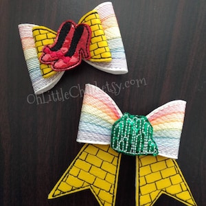 May include: Two handmade hair bows with a Wizard of Oz theme. One bow features red sequined ruby slippers, and the other has a green Emerald City design. Both bows have yellow brick road and rainbow accents.