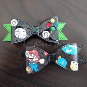 May include: Two black and green fabric hair bows with a video game theme. One bow features a black and white video game controller with green, red, and yellow buttons. The other bow features a cartoon image of Mario from the Super Mario video game series.