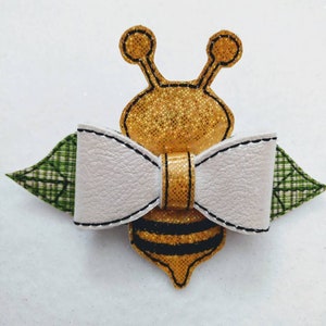 May include: A whimsical bee-shaped hair bow. The bee features a gold glitter body with black stripes, a white bow, and green plaid wings. The edges are outlined with black stitching, creating a charming accessory.