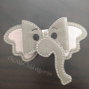 May include: A gray elephant-shaped hair bow with a textured appearance. The bow features a light pink inner ear and is outlined with white stitching. The elephant has black eyes and a trunk. The text "OhLittleChild.etsy.com" is visible.